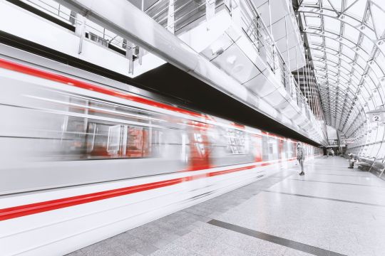 Ein weißer Zug mit roten Streifen fährt schnell durch einen Bahnhof mit Glas-Tonnendach, es ist nicht zu erkennen, dass es sich um die überirdische Prager Metro-Endstation Černý Most handelt, die nach dem gleichnamigen Plattenbau-Stadtteil benannt ist.