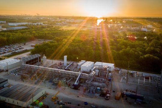 Kraftwerksanlage in einem großen Industriegebiet in flacher Landschaft mit Bäumen zwischen den Anlagen, aufgenommen im Gegenlicht.