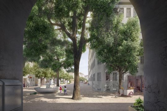 Durch einen Torbogen schauen wir auf eine ebene sandige Fläche zwischen Häusern, in der Mitte ein Brunnen, an dem Kinder mit Gießkannen spielen. Große Bäume stehen zwischen den Häusern, ein Lastenrad vor einem Laden, in dem Menschen einkaufen. Viele Fahrr