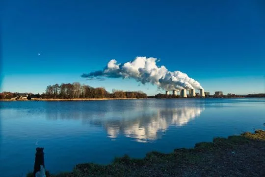 Braunkohlekraftwerk Jänschwalde mit weiß dampfenden Kühltürmen hinter einem See, Wasser und Himmel sind tiefblau.