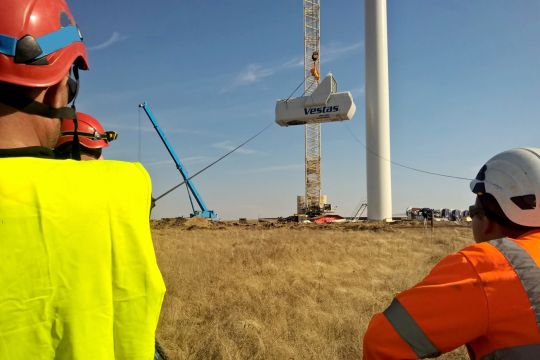 Ein Kran hebt eine Windturbine mit der Aufschrift Vestas neben einem Windrad-Turm in die Höhe, im Vordergrund schauen drei Arbeiter in Montur und mit Helm zu.