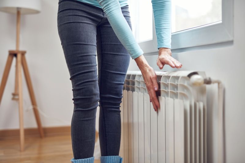 Eine Frau in dicken Socken hält die Hände dicht an einen Heizkörper, um sich zu wärmen.