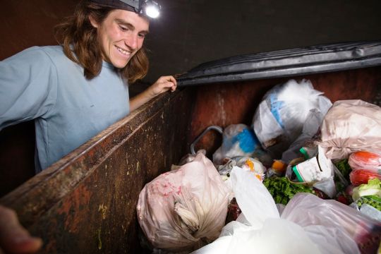 Ein junger Mann mit Stirnlampe freut sich über die Lebensmittel, die er in einem Müllcontainer findet, den er gerade geöffnet hat.