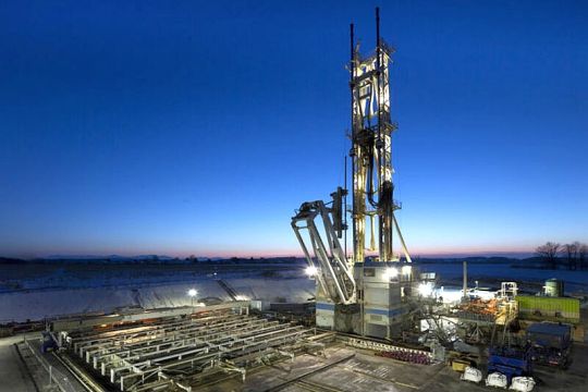 Senkrecht aufragende Bohranlage mit Nebenanlagen auf einem Bauplatz vor tiefblauem Abendhimmel.