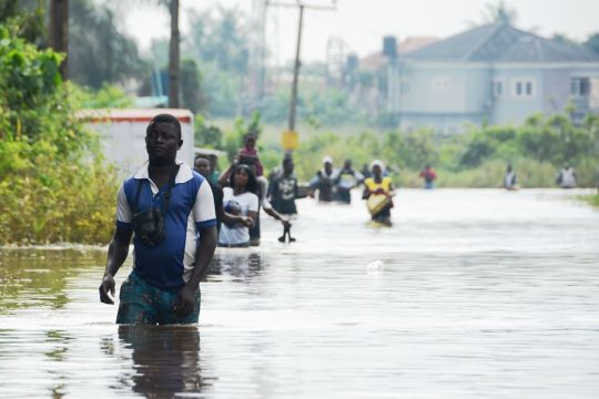 Menschen gehen durch hüfthohes Wasser, im Hintergrund ein neues hellblaues Wohnhaus. Flutkatastrophe im südwest-nigerianischen Bundesstaat Ogun im Oktober 2019.