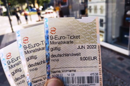 Hand hält drei Neun-Euro-Tickets für Juni, Juli und August 2022, im Hintergrund eine Straßenszene.