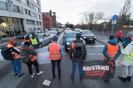 Ein halbes Dutzend Menschen in Warnwesten blockiert eine mehrspurige Straße, auf ihrem Transparent steht: Aufstand der Letzten Generation.