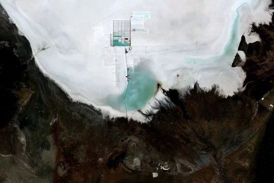 Luftaufnahme: Weißer Salzsee mit ausfransenden Ufern in dunkelbrauner Landschaft, darin türkisfarbene Flächen sowie eine rechteckige Struktur - dort findet die Lithiumgewinnung statt.