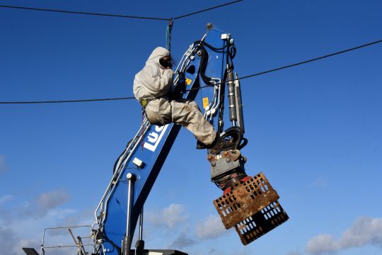 Eine Person in grauem Schutzanzug hängt an einem Seil, hinter ihr der blaue Greifarm einer Baumaschine.