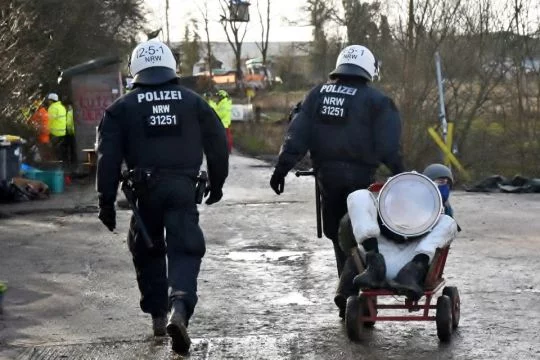 Zwei behelmte Polizisten fahren einen Aktivisten mit einem Bollerwagen weg.