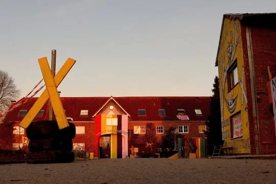 Zweistöckiges Backsteinhaus auf einem ehemaligen Bauernhof, der Torgiebel ist mit Regenbogen-Farben bemalt. Vor dem Haus steht ein großes gelbes X aus Holz.