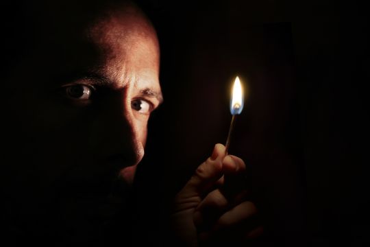 Ein Mann hält in der Dunkelheit ein brennendes Streichholz hoch und schaut dich ängstlich an.