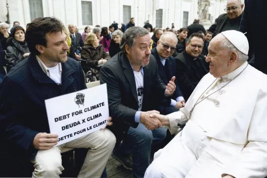 Papst Franziskus schüttelt Juan Pablo Olsson die Hand. Daneben sitzt Esteban Servat mit einem Schild: Debt for Climate.