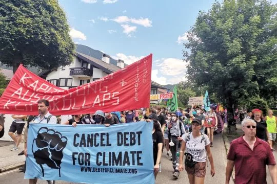 Demonstrationszug in einer beschaulichen Straße, auf einem Transparent steht: Schuldenerlass für Klimaschutz. Auf einem anderen Transparent steht: Kapitalismus ist Ausbeutung plus Klimakollaps.