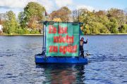 Ein kleiner motorisierter Ponton schwimmt auf der Spree, darauf ein winkender Mann und ein großes grün-rotes Banner: Es ist noch nicht zu spät!