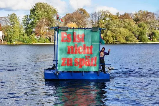Ein kleiner motorisierter Ponton schwimmt auf der Spree, darauf ein winkender Mann und ein großes grün-rotes Banner: Es ist noch nicht zu spät!