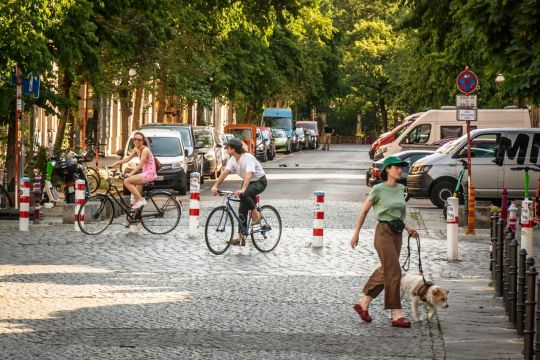 Eine Kreuzung in einem Berliner Wohngebiet ist durch eine Reihe Poller diagonal gesperrt, Menschen bewegen sich per Rad und zu Fuß hindurch, während der Autoverkehr auf Anwohnerverkehr beschränkt bleibt.