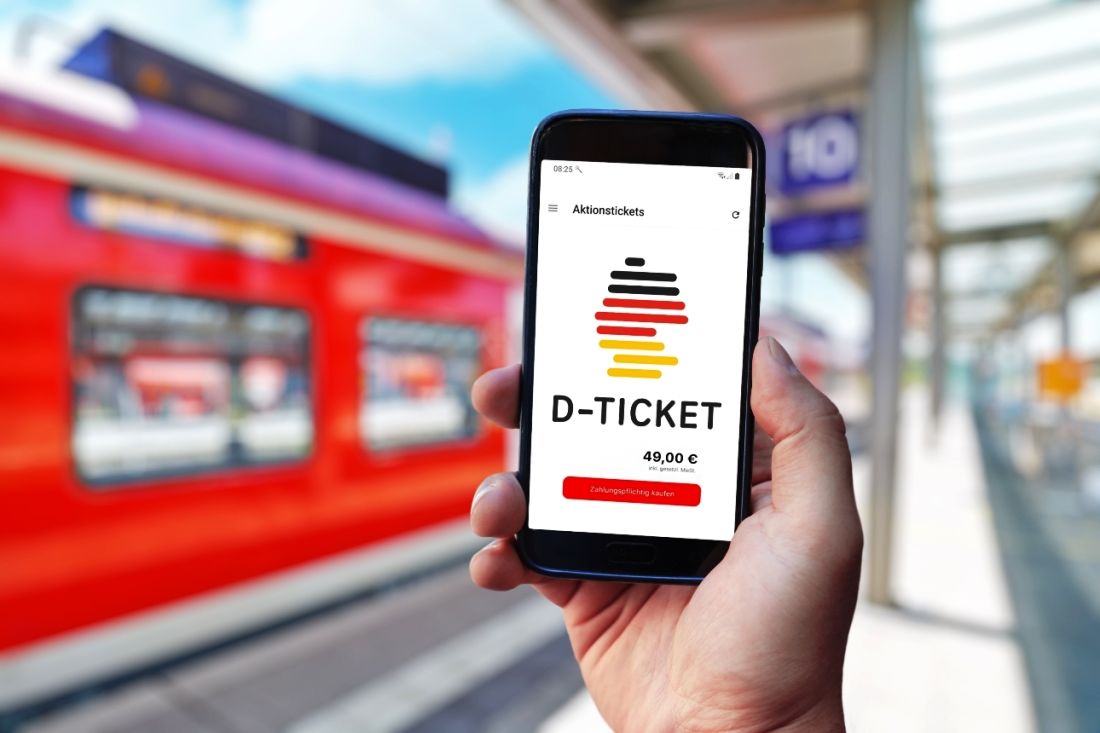 Eine Hand hält ein Handy, auf dem die D-Ticket-App zu sehen ist, im Hintergrund steht ein Regionalzug an einem Bahnsteig in Dossenheim bei Heidelberg.