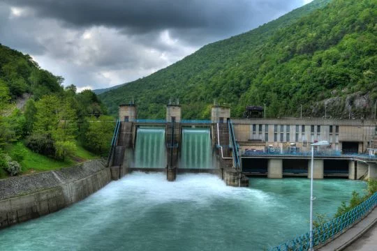 Ein Wasserkraftwerk vor einer grünen Bergkulisse. Das Wasser fließt neben dem Kraftwerk über den Staudamm.