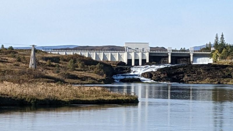 Wasserkraftwerk aus hellem Beton in einer hügeligen, kargen Landschaft, vom Ablauf und etwas von unten her gesehen.