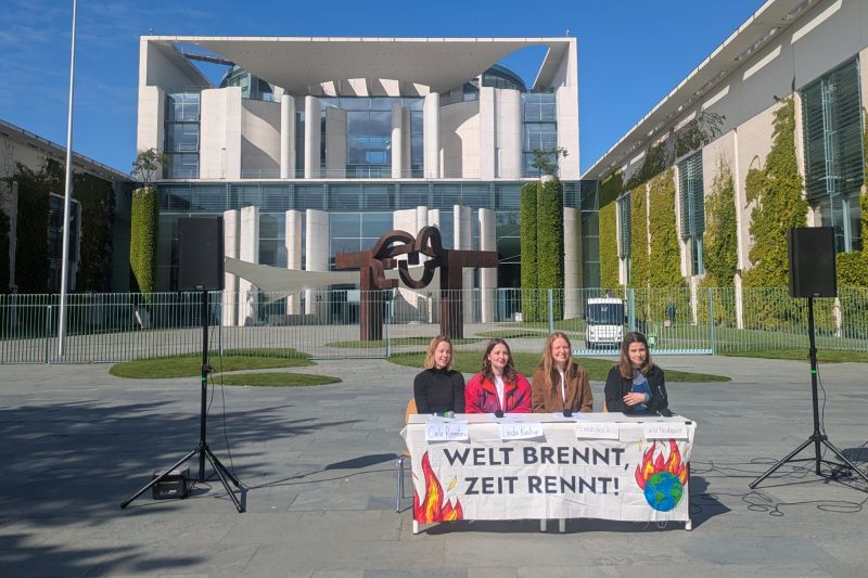 Vier Frauen sitzen hinter einem improvisierten Podium vor dem Bundeskanzleramt in Berlin, eine spricht ins Mikro. Auf einem Transparent vor ihnen steht: Welt brennt, Zeit rennt!