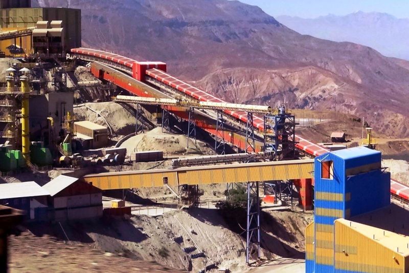 Umfangreiche Industrieanlagen mit eingehausten Förderbändern und mehrstöckigen Industriehallen, alles in einer vegetationslosen Hochgebirgslandschaft.