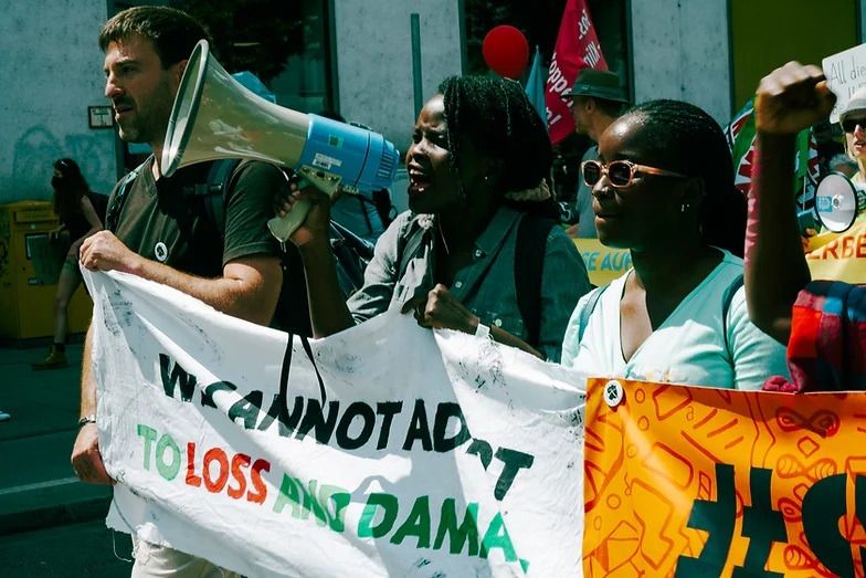 Zwei afrikanische Frauen an der Spitze einer Demonstration mit Transparenten und einem Megafon.