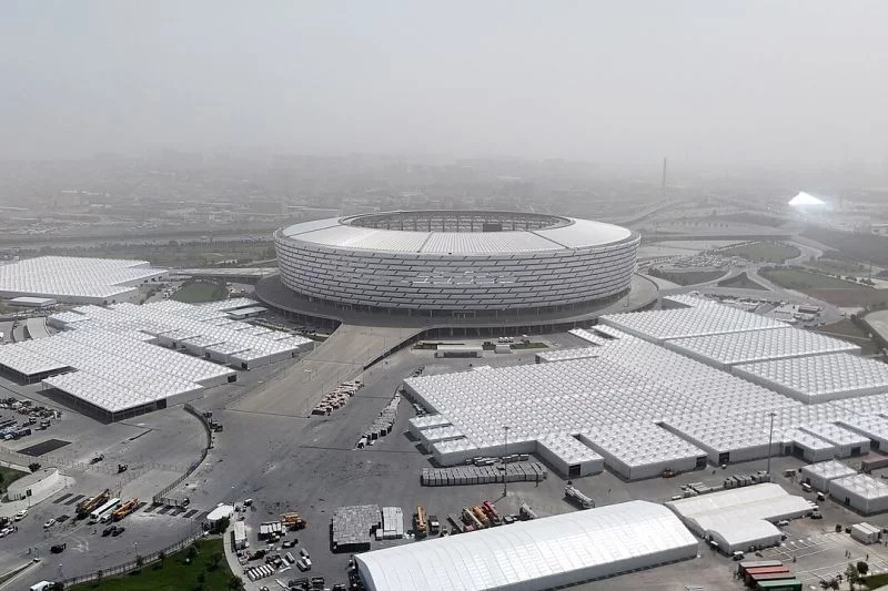 Luftaufnahme: Ein rundes Stadion, daneben drei große Veranstaltungshallen-Komplexe, alles in Weiß, dahinter die Stadt im Nebel.