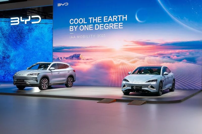 Messepräsentation des chinesischen Autokonzerns BYD mit zwei Autos und einem futuristischen Hintergrund in hellblau und orange mit der englischen Aufschrift: Die Erde um ein Grad abkühlen.