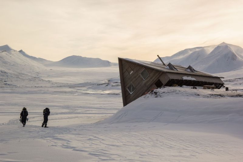 In einer Schneelandschaft ist eine Holzhütte auf einem Hügel ins Kippen geraten, zwei Menschen schauen sich das an.