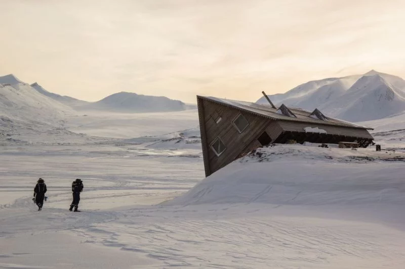 In einer Schneelandschaft ist eine Holzhütte auf einem Hügel ins Kippen geraten, zwei Menschen schauen sich das an.
