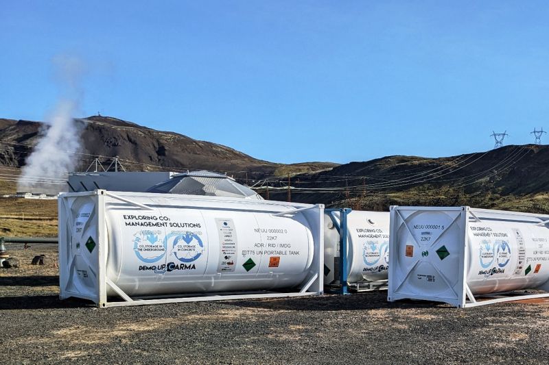 Weiße Gasbehälter in Container-Rahmen mit der Aufschrift Demoupcarma stehen in einer kargen, bergigen Landschaft.
