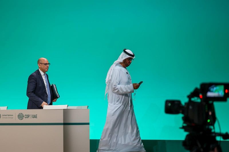 Sultan Al Jaber geht von der Bühne der Klimakonferenz, während er auf sein Handy schaut, hinter ihm geht Simon Stiell. 