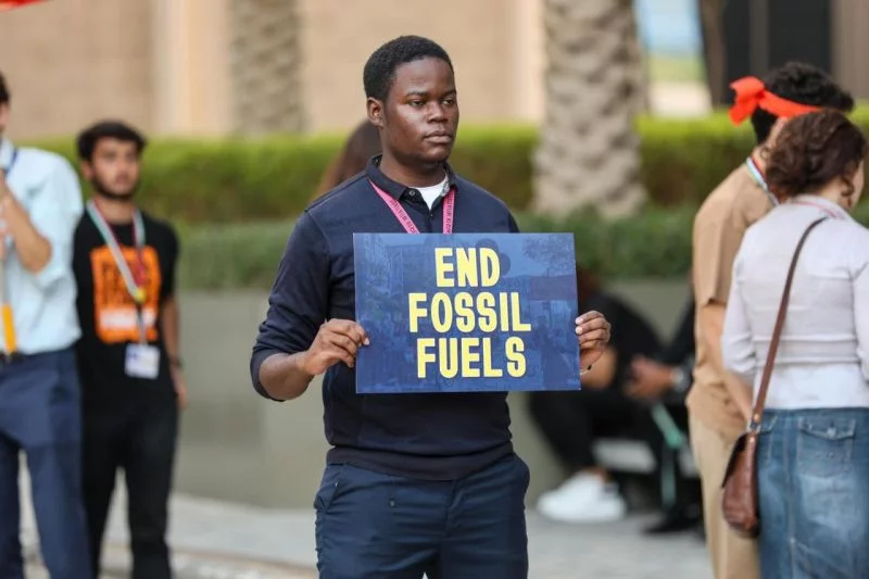 Auf dem Gelände der Klimakonferent in Dubai steht zwischen anderen Menschen ein Afrikaner und hält ein einfaches Schild hoch, darauf steht auf Englisch: Schluss mit fossilen Energien.