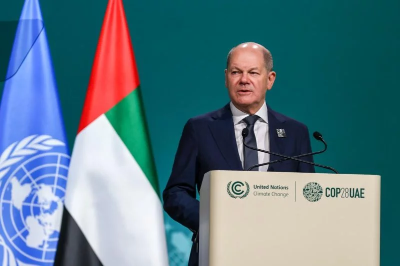 Bundeskanzler Olaf Scholz spricht am Pult des Klimagipfels COP 28, neben ihm die Flaggen der Emirate und der UN.