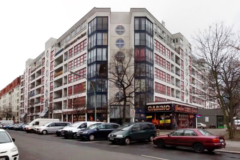 Achtstöckiges Eckhaus in Plattenbauweise, einige Fenster sind rund oder als großzügige Eckfenster ausgeführt, unten sind Läden, vor denen Autos geparkt sind.