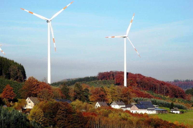 In bewaldetem, herbstlich eingefärbtem Hügelland stehen neben einem Dörfchen zwei große, weiße Windräder.