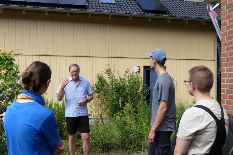 Ein Mann gestikuliert, Jugendliche hören zu, alle stehen, dahinter eine gelbe Einfamilienhauswand.