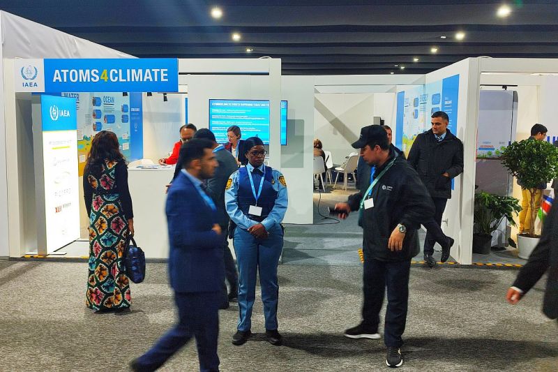 Einige Menschen an improvisiert wirkenden Pavillons in einer Konferenzhalle, auf einem Schild steht ein Slogan der Internationalen Atomenergiebehörde IAEA: Atoms for Climate.