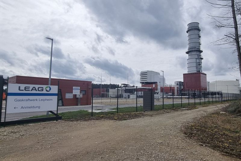Ein modernes Gaskraftwerk mit einem Schornstein, schräg vom Werkstor aus gesehen, auf einem Schild steht der Name des Eigentümers Leag.