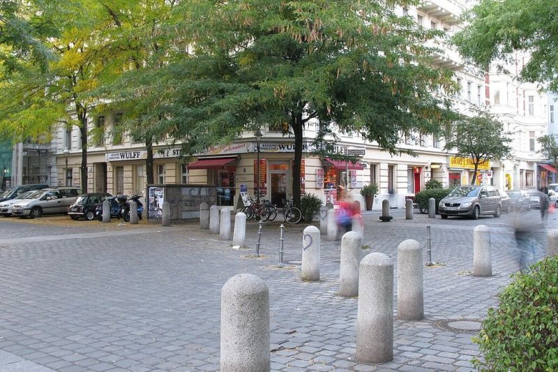 Eine Reihe aus Steinpollern läuft diagonal über die gepflasterte Straßenkreuzung in dem Viertel mit Gründerzeithäusern. 