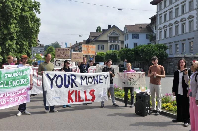 Hilda Arrieta spricht ins Mikro, daneben eine kleine Demonstration mit handgemalten Transparenten wie: Euer Geld tötet! Kein Steuergeld für Glencore!