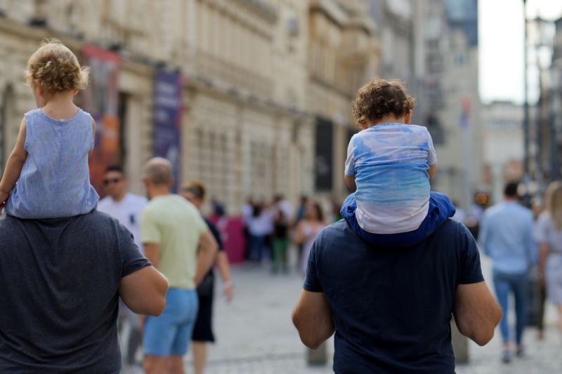 Zwei Väter mit Kleinkindern auf ihren Schultern in einer Fußgängerzone.