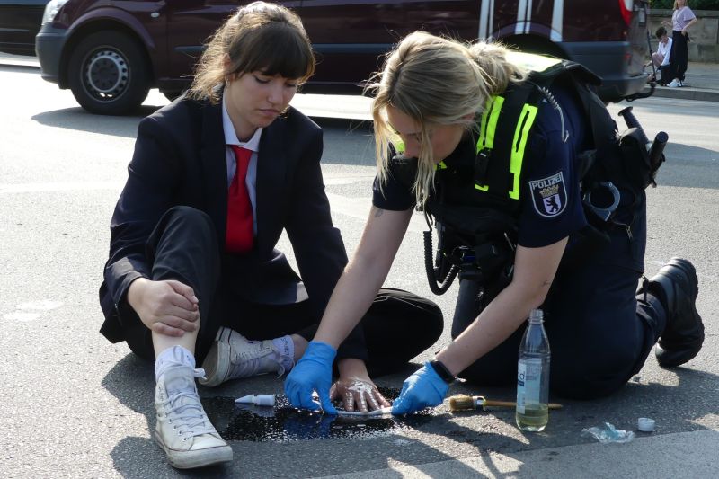 Carla Hinrichs sitzt im Anzug auf der Straße, an der sie ihre Hand festgeklebt hat, neben ihr kniet eine Polizistin mit blauen Handschuhen und versucht den Kleber zu lösen.