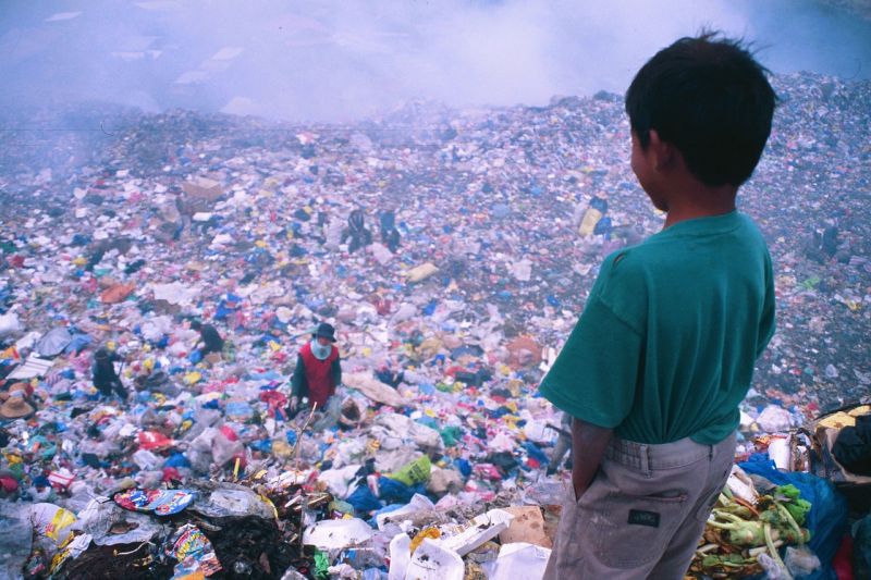 Einige Menschen sammeln Brauchbares auf einer Mülldeponie auf den Philippinen, im Vordergrund steht ein Kind am Rand der Deponie und schaut auf die Sammelnden herunter.