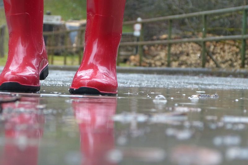 Eine Person steht im Regen, man sieht nur rote Gummistiefel und einen nassen, glatten Boden.