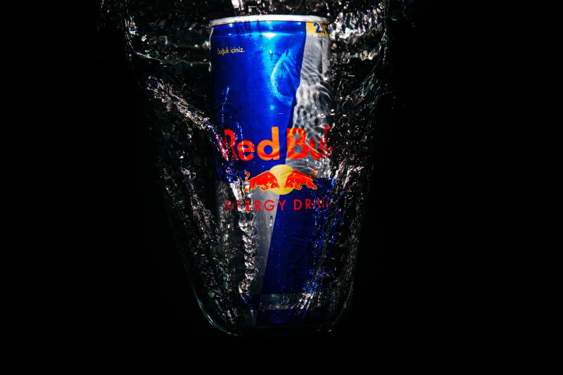 Um eine Dose Red Bull spritzt Wasser. Alles vor schwarzem Hintergrund.
