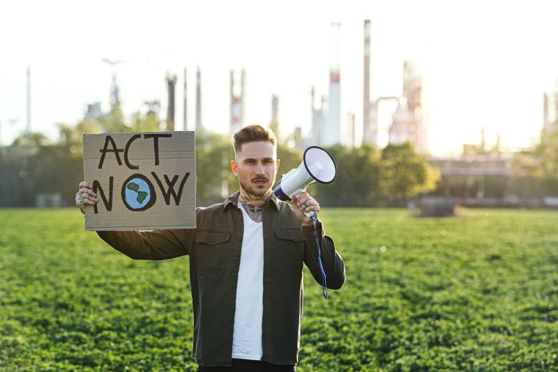 Ein junger Aktivist steht mit einem Megafon auf einer Wiese, weit im Hintergrund ist ein Industriebetrieb nur undeutlich zu erkennen, auf seinem Plakat steht auf Englisch: Handelt jetzt!