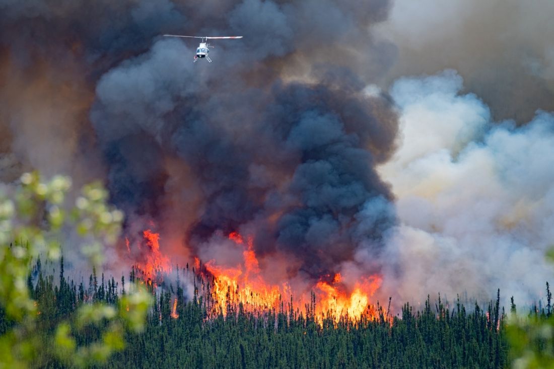 Groißflächig brennender Wald in Kanada, vor dichten dunkelgrauen Rauchschwaden ist ein kleiner Hubschrauber zu sehen.