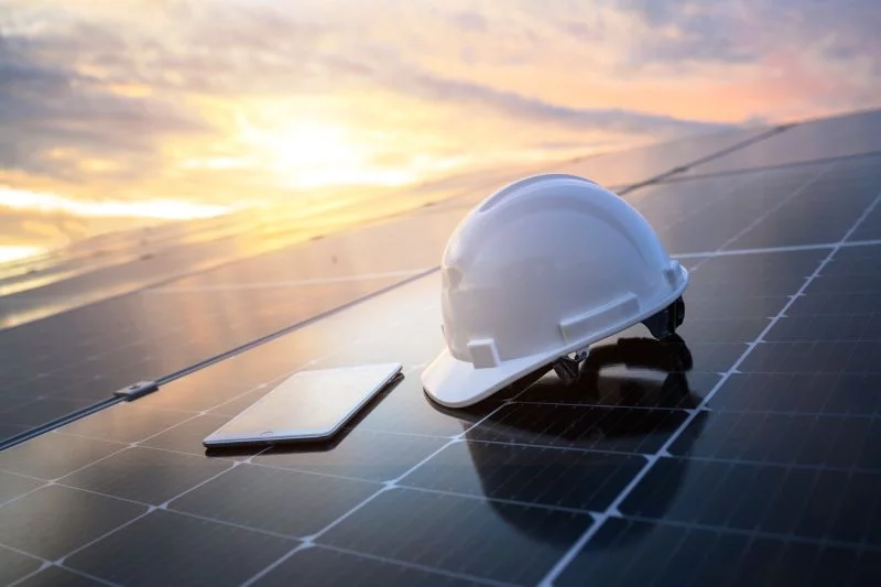 Ein weißer Schutzhelm und ein Tablet liegen auf einem Dach, das großflächig mit Solarpaneelen bedeckt ist.